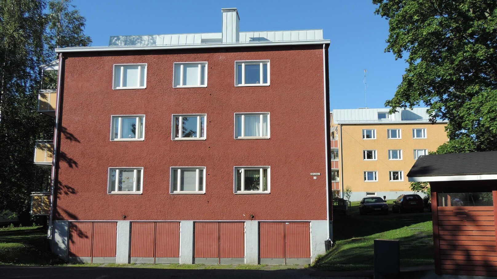 Etualalla punainen rapattu vanha kerrostalo, sokkelissa neljä autotallia punaisine ovineen, harmaa peltikatto, taustalla samanlainen keltainen talo, etualalla varjossa parkkipaikkaa